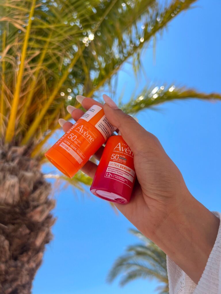 Ruka drži Avène Ultra Stick SPF50 i Avène Ultra Serum SPF50 ispod palme na sunčanom danu
