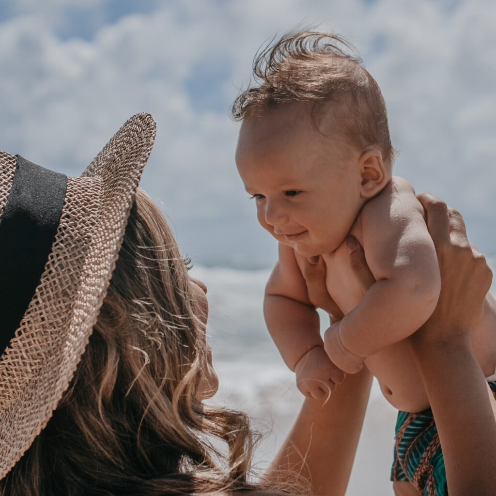 Žena sa šeširom podiže bebu prema suncu na plaži, uz more i ljetno nebo u pozadini