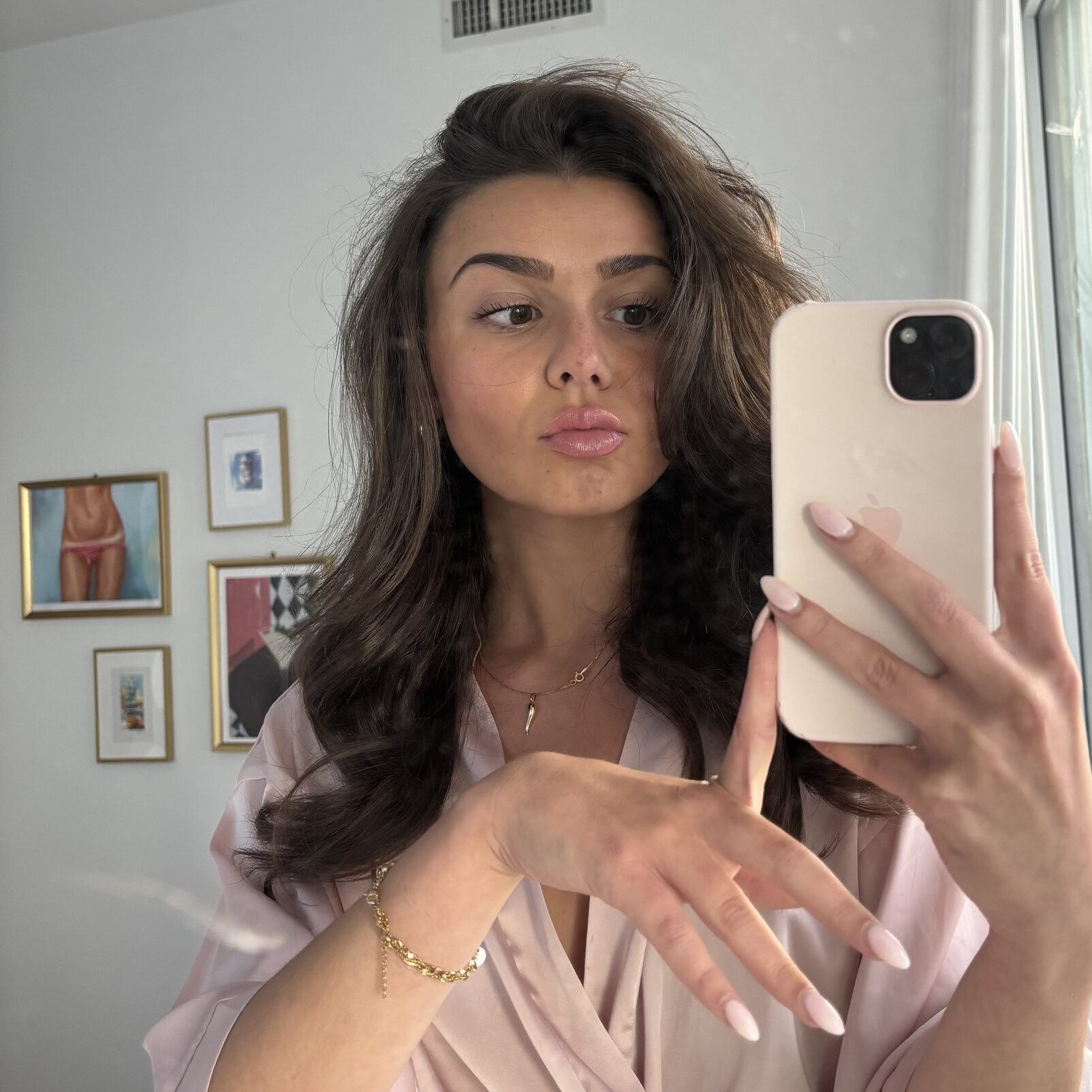Mlada žena s dugom smeđom kosom radi selfie u ogledalu, noseći ružičasti satenski ogrtač i držeći telefon