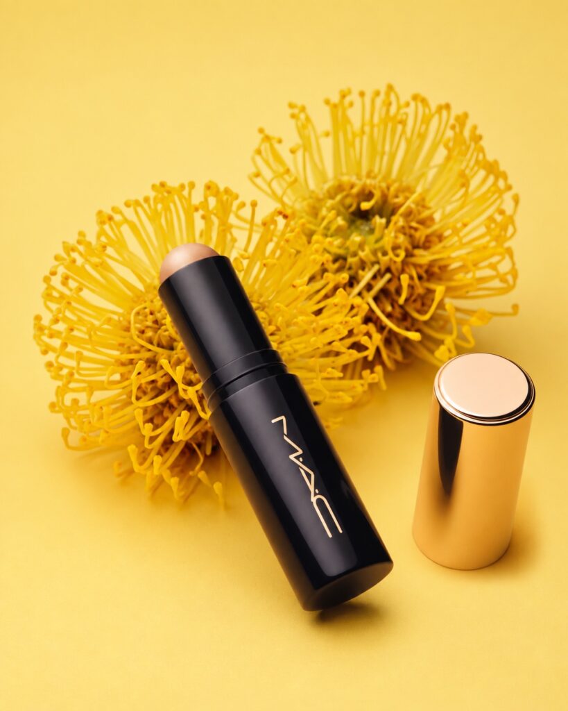 Otvoreni M·A·C Studio Radiance Skin Tint Stick uz žute cvjetove i zlatni poklopac
